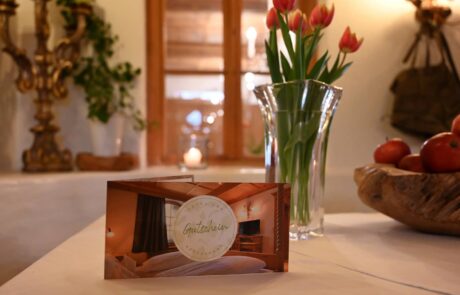 Ein Gutschein auf einem Tisch im Hotel, umgeben von Blumen, Äpfeln und gemütlicher Einrichtung für einen entspannten Aufenthalt.