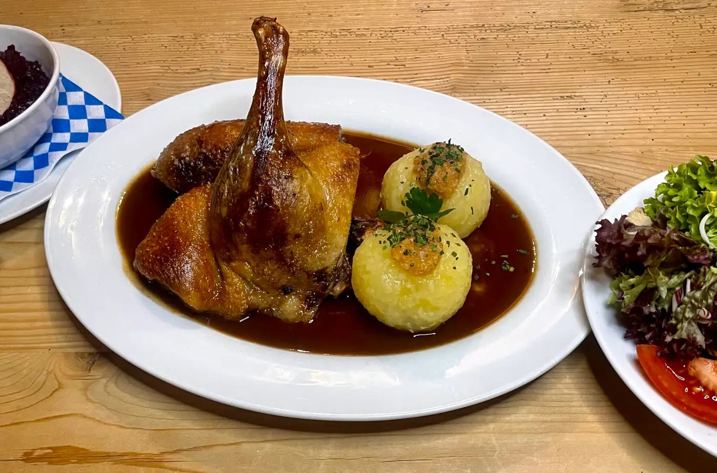 Ein Teller mit knuspriger Hähnchenkeule, zwei Kartoffelknödeln und einem frischen Salat auf rustikalem Holztisch.