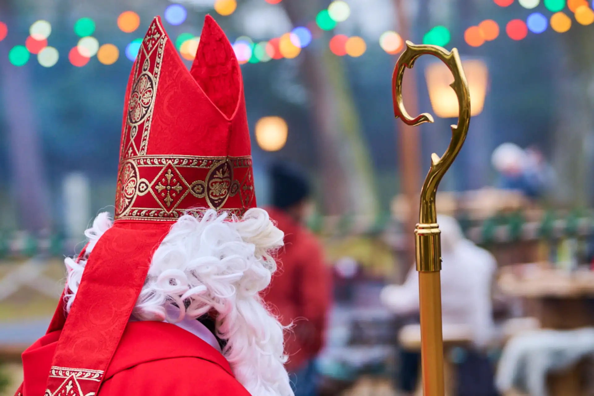 Der Nikolaus in rotem Gewand mit goldenem Stab steht auf einer festlichen Weihnachtsfeier, umgeben von bunten Lichtern und Gästen.