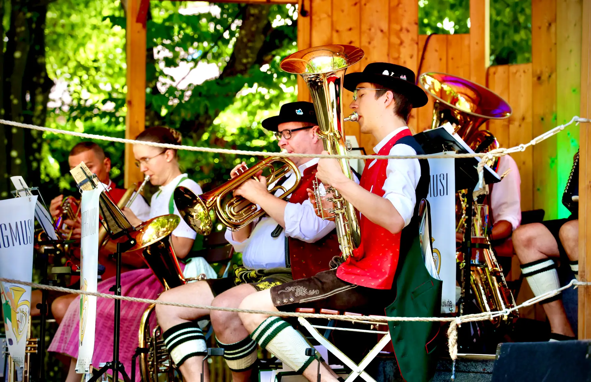 Ein Blasmusikensemble spielt auf einer Bühne bei einem bayerischen Volksfest, umgeben von grüner Natur und Holzbau.