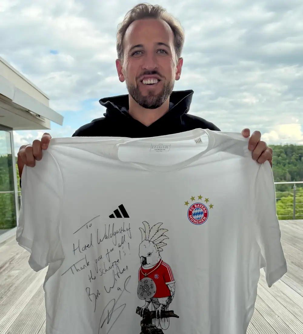 Ein Mann mit einem T-Shirt des FC Bayern München, signiert und als Dankeschön für das Hotel Waldgasthof, steht auf einer Terrasse.